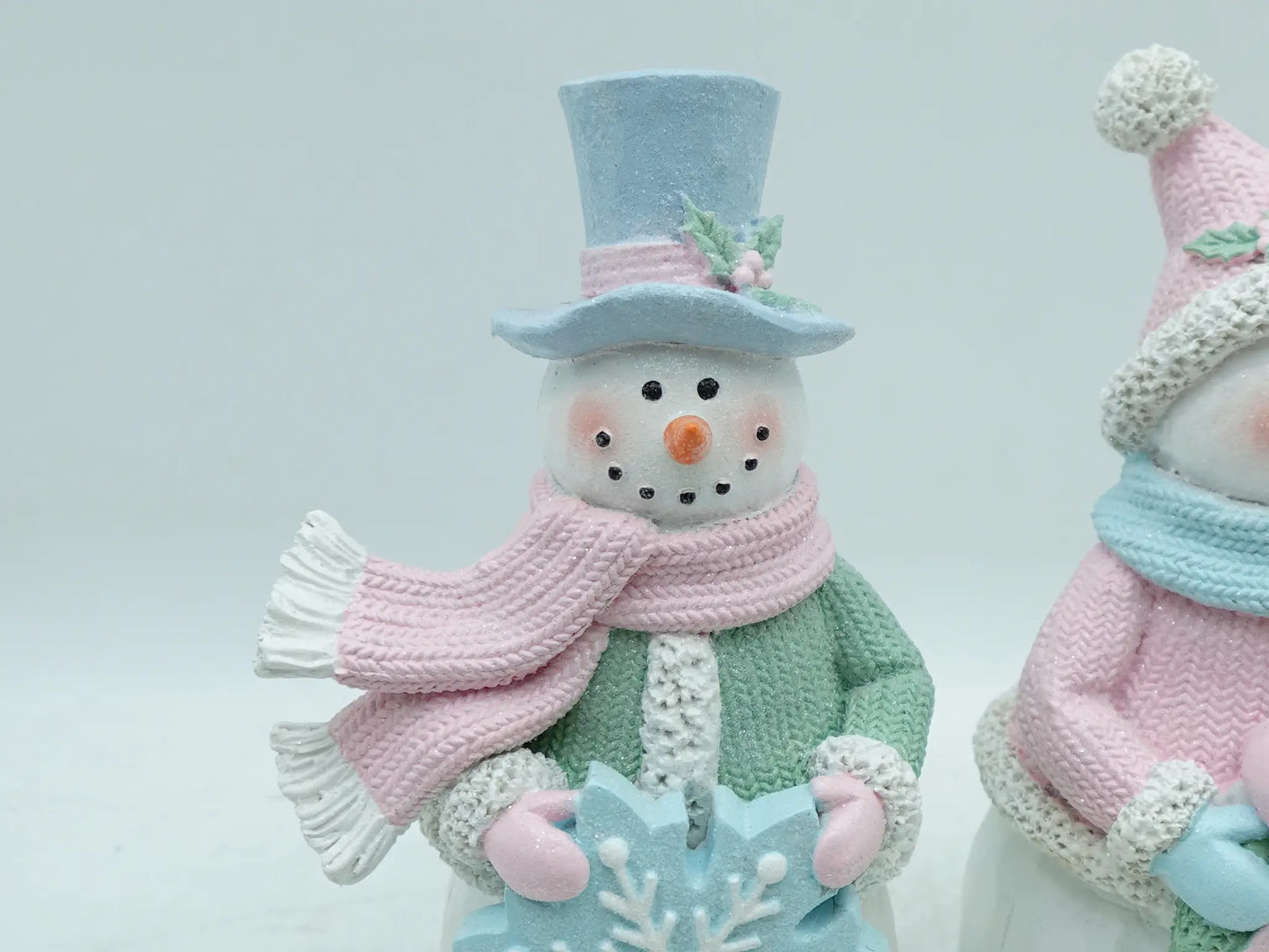 Close-up of male snowman figurine in sky-blue top hat, pink scarf, and mint-green coat holding an icy-blue snowflake