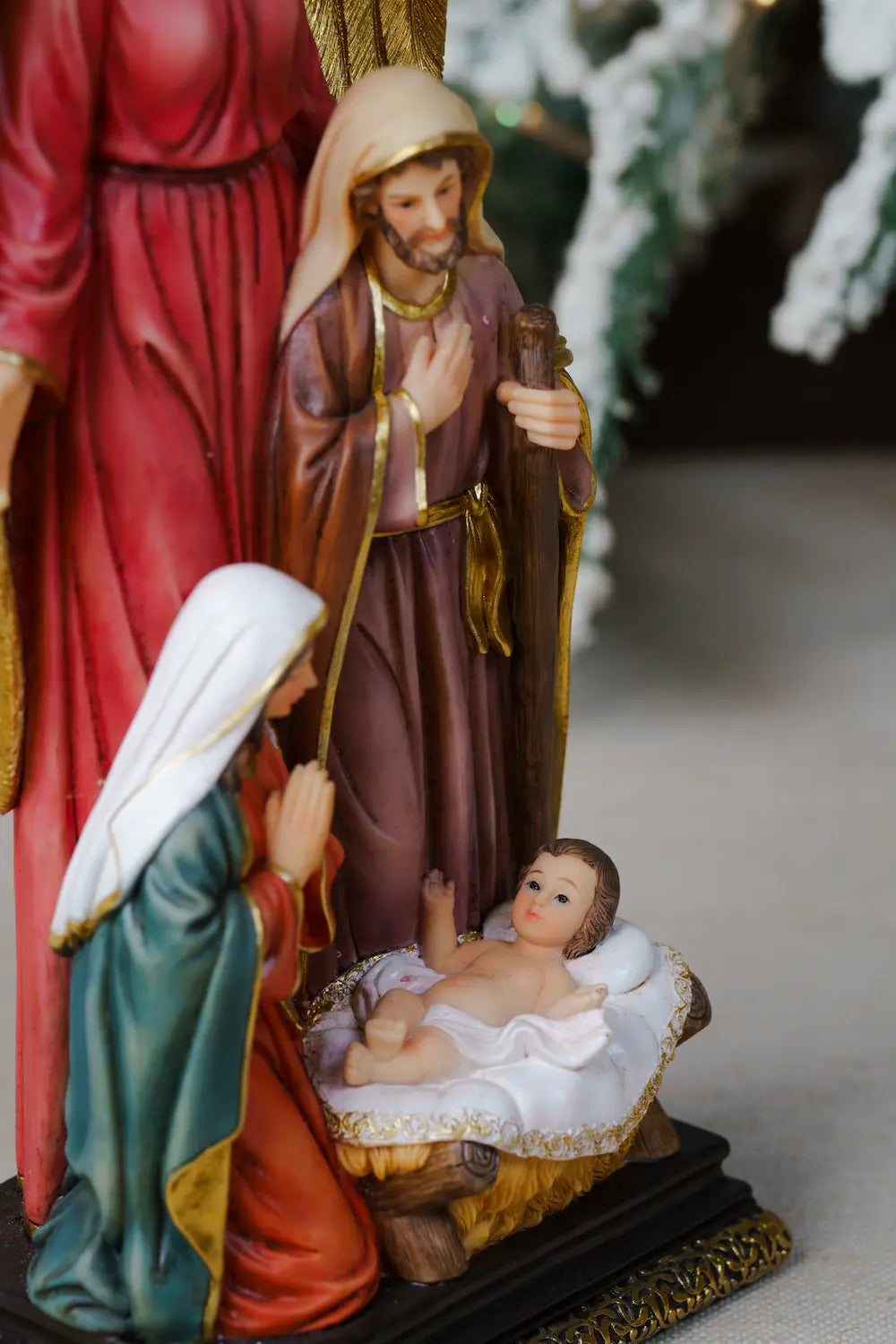 A close-up of nativity figurines: Joseph standing, Mary kneeling in prayer, Baby Jesus in the manger, with a large gold-winged angel behind them.