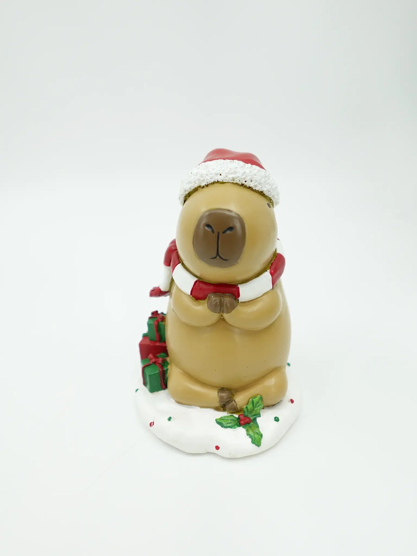 A resin Christmas capybara figurine wearing a Santa hat and scarf, sitting on a snowy base with small wrapped gifts.