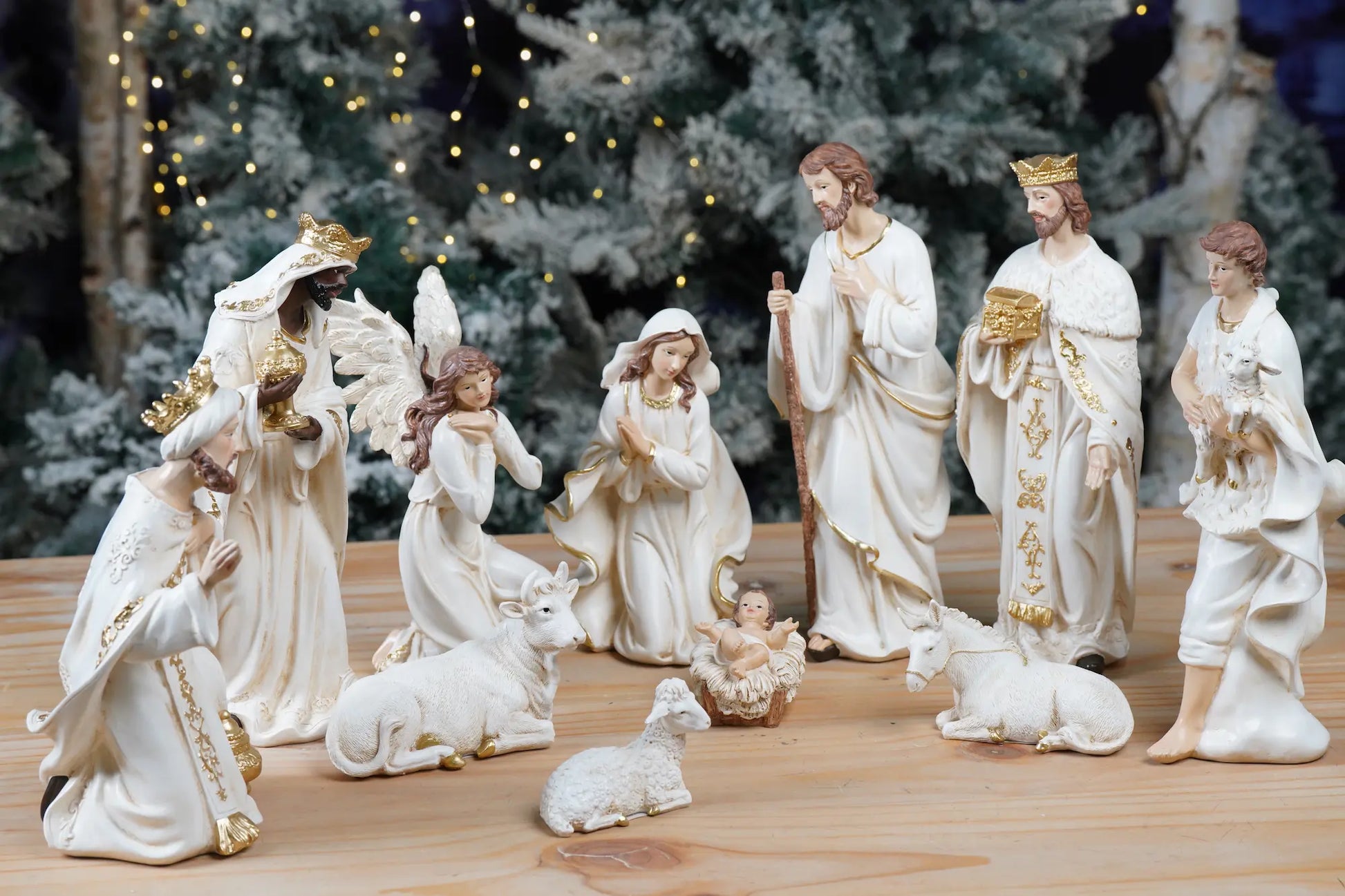 Full white and gold nativity set arranged on pine wood surface with frosted Christmas tree background