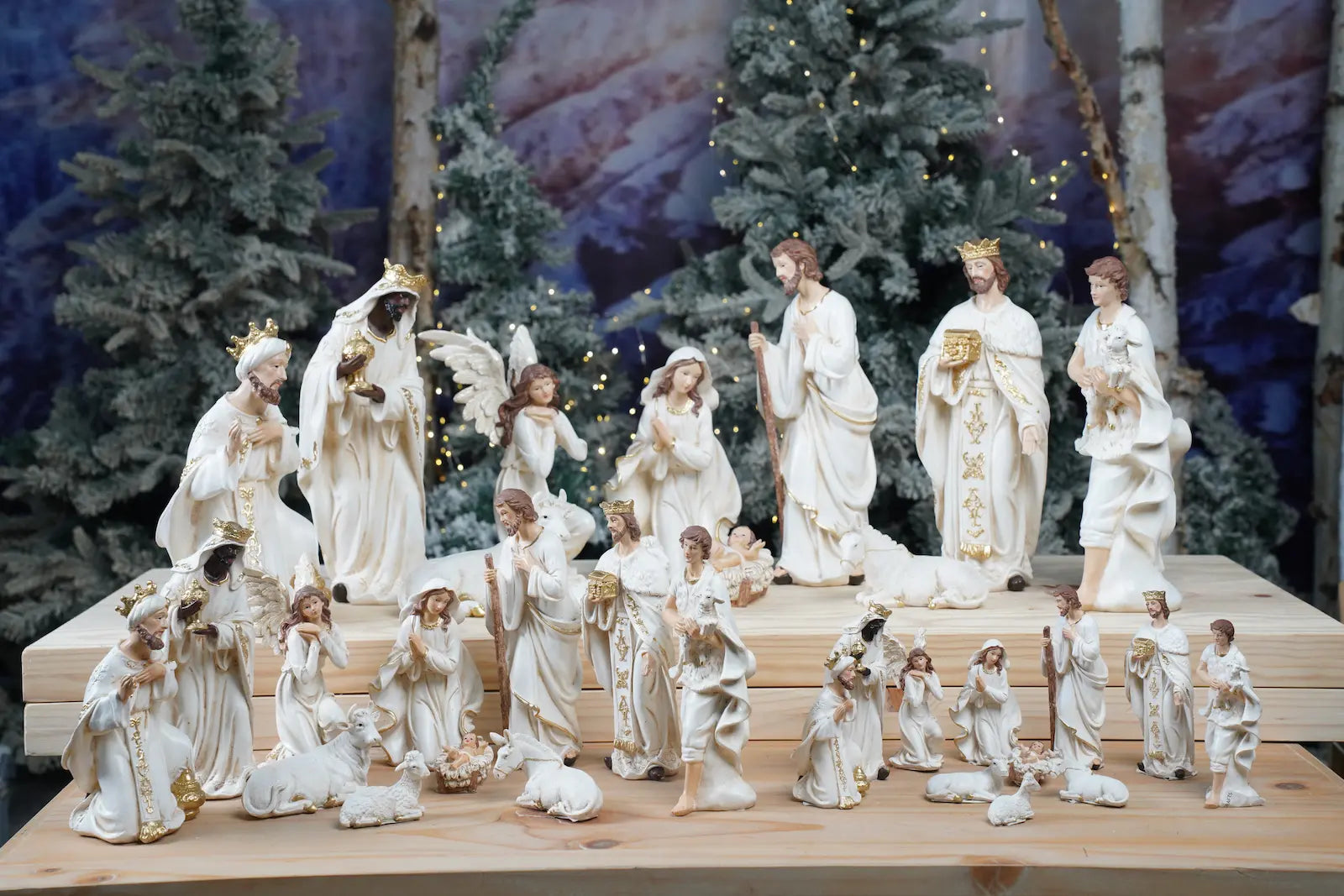 Multiple sizes of white and gold nativity sets displayed together on tiered wooden platform with winter tree backdrop