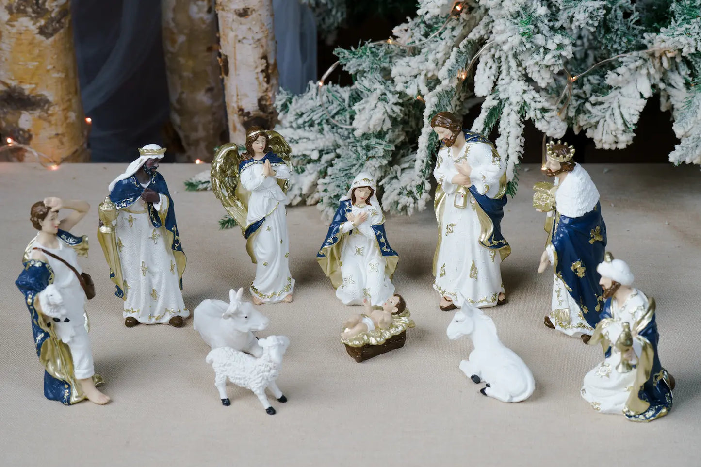 Christmas nativity figurines arranged on a tabletop in front of a flocked Christmas tree.
