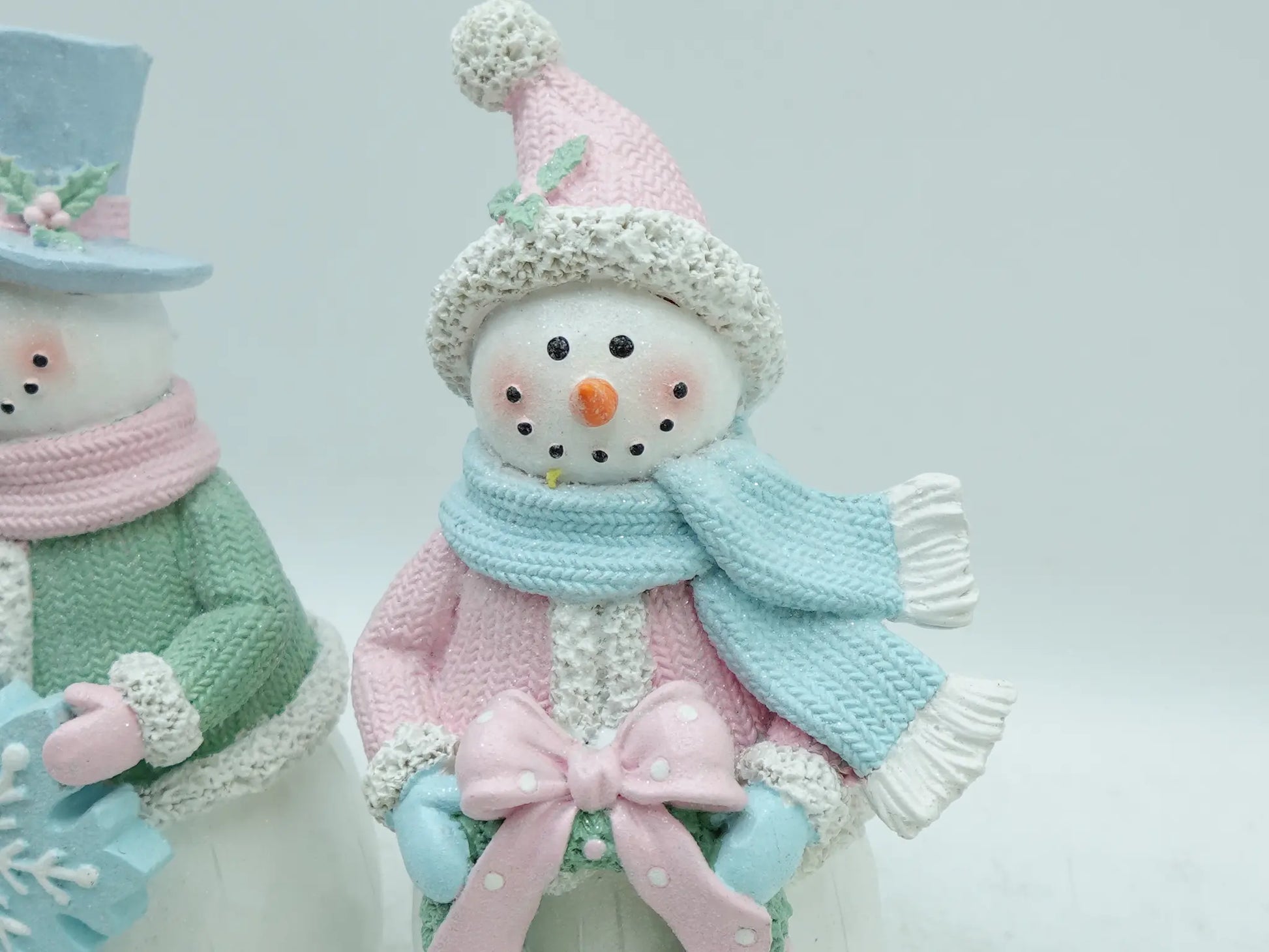 A close-up of a knitted snowman figurine wearing a pink hat with a pom-pom, a light blue scarf, and a pink bow, with part of another snowman visible on the left.