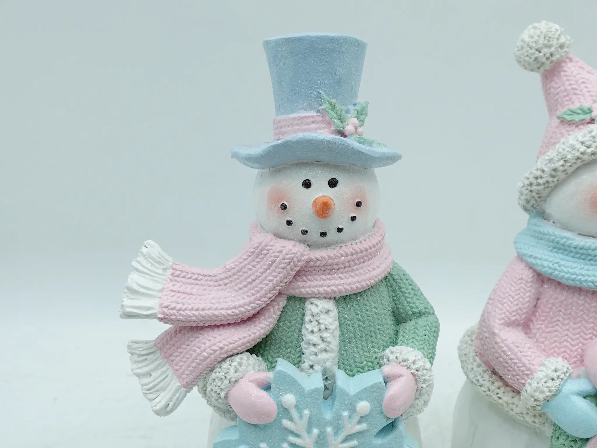 Close-up of male snowman figurine in sky-blue top hat, pink scarf, and mint-green coat holding an icy-blue snowflake