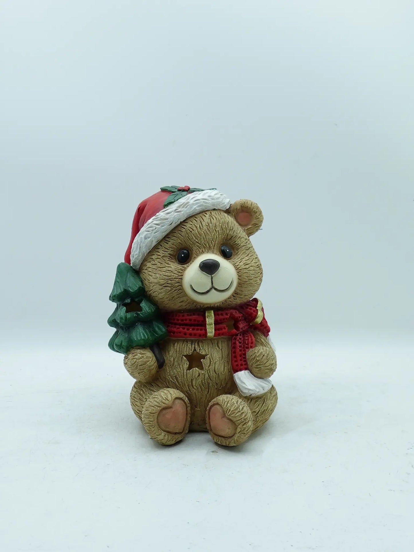 A holiday teddy bear figurine carrying a green Christmas tree, photographed against a plain light gray background.