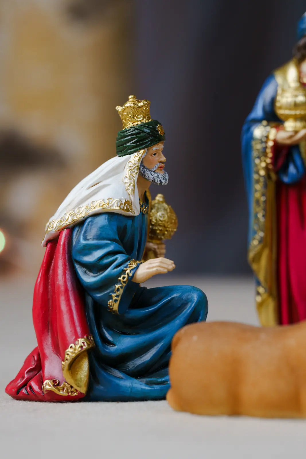Nativity figurine of a kneeling king in blue, red, green, and gold robes, with part of another regal figurine visible in the background.