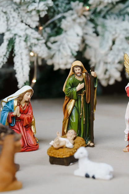 A set of hand-painted Christmas nativity figurines arranged on a neutral surface. The figurines include Mary in red and blue, Joseph in green and gold, baby Jesus in a brown manger.