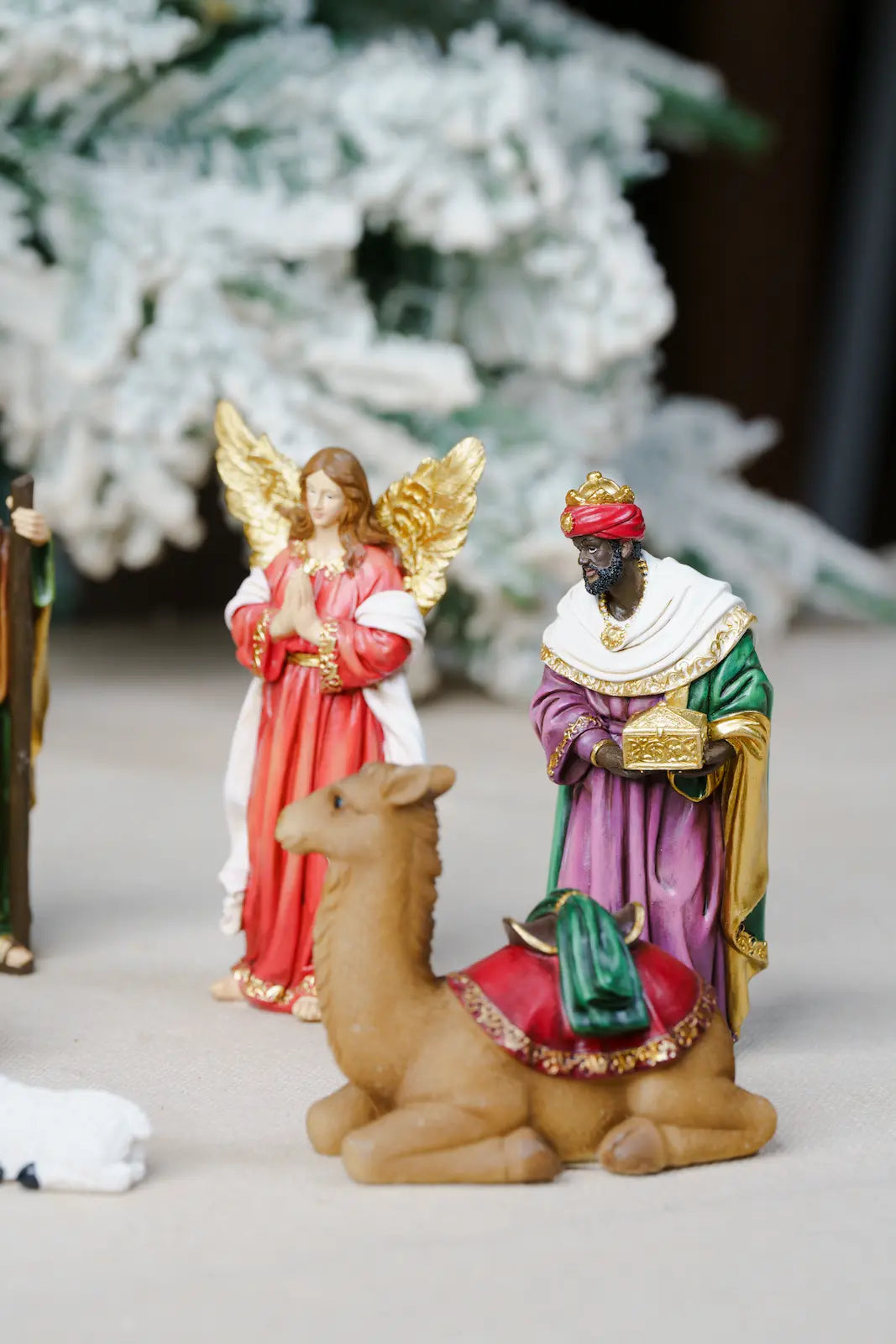 small nativity figurines set on a light surface, featuring an angel in red and white robes with gold wings, a wise man in purple and green garments, and a camel in tan with a red and green saddle.