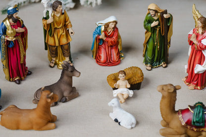 A set of colorful Christmas nativity figurines including Mary, Joseph, baby Jesus, animals, and wise men, arranged on a neutral beige surface.