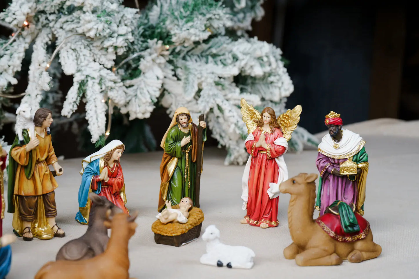 Christmas nativity set figurines arranged on a tabletop in front of a flocked Christmas tree. 