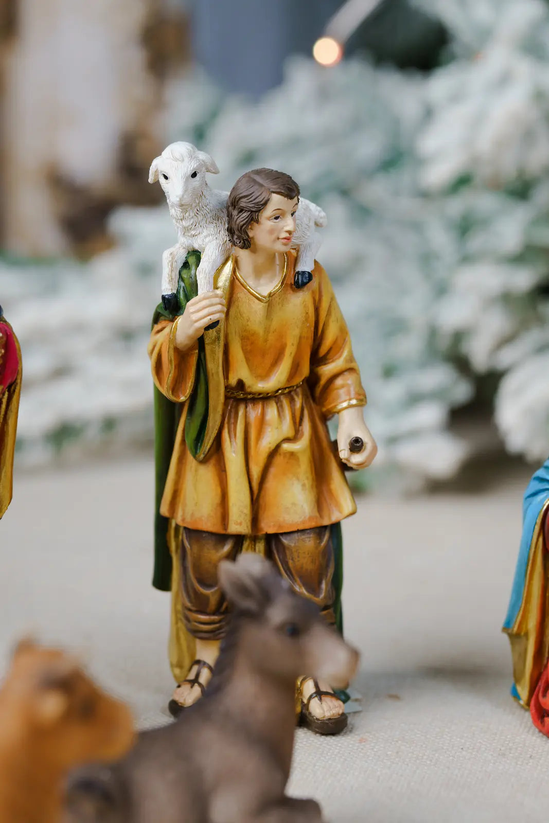 A hand-painted nativity figurine of a shepherd carrying a white sheep on his shoulders, wearing a golden-brown tunic and green cloak.