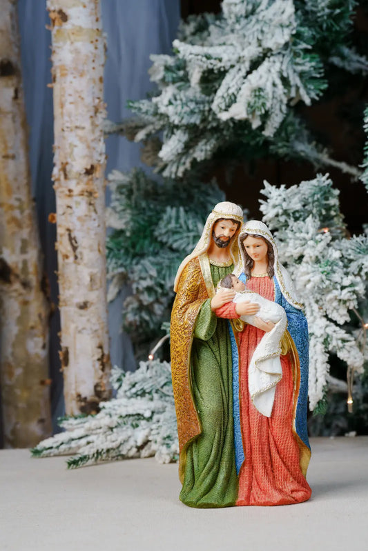 A Christmas nativity figurine of Mary, Joseph, and baby Jesus, styled in front of a snowy Christmas tree and birch trees.