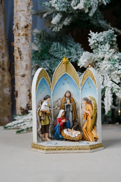 A tabletop nativity scene with three arched panels showing the Holy Family, angels, and baby Jesus in a gold-tray base.