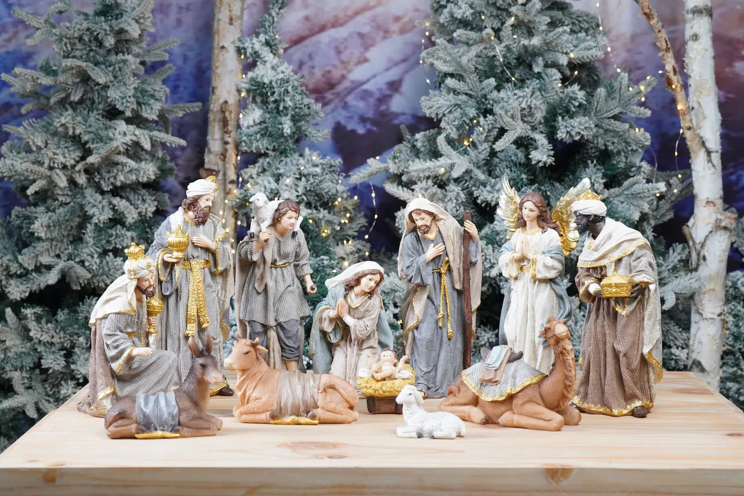 Full 12-piece grey and gold nativity set arranged on pine wood surface with frosted winter tree backdrop