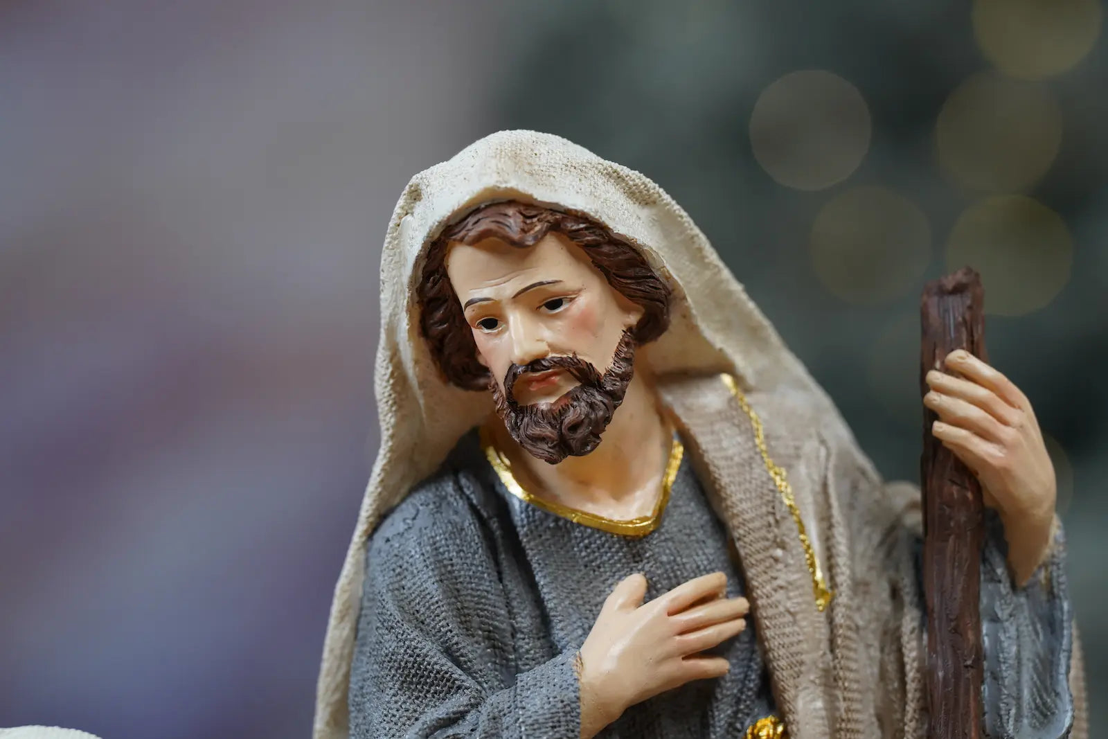Joseph nativity figurine close-up — grey textile-texture robes with gold trim and hand-over-heart devotional pose