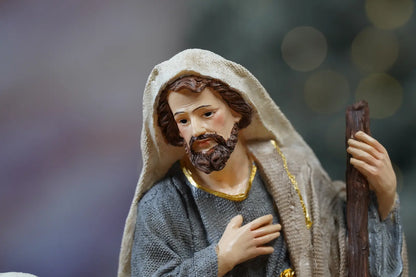 Joseph nativity figurine close-up — grey textile-texture robes with gold trim and hand-over-heart devotional pose
