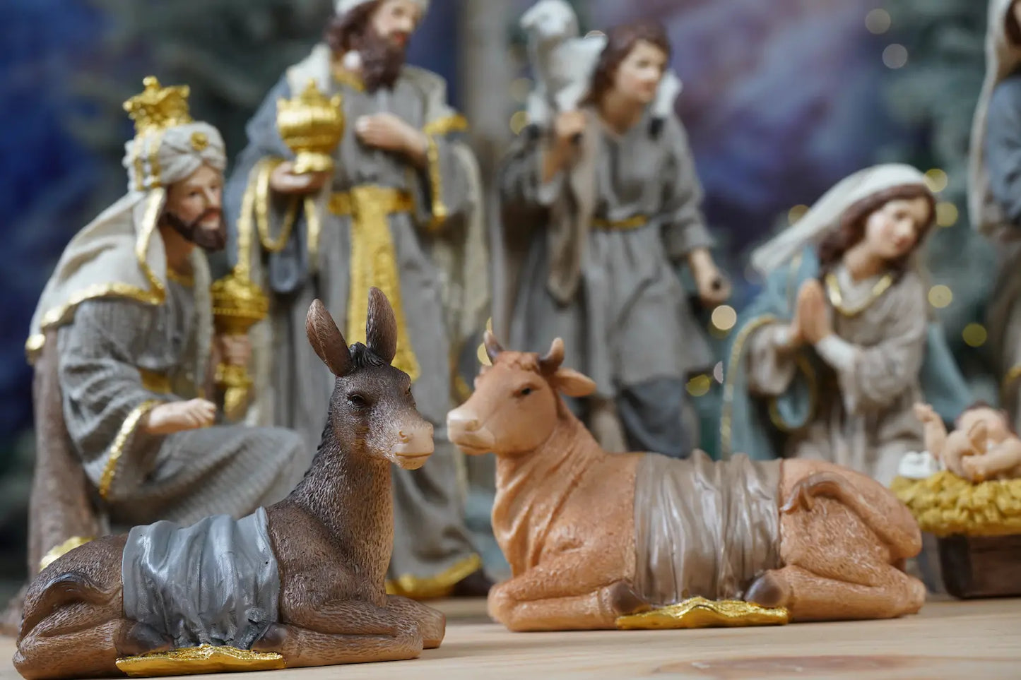 Nativity stable animals close-up — donkey with grey fabric drape and ox resting beside Baby Jesus manger with figures in background