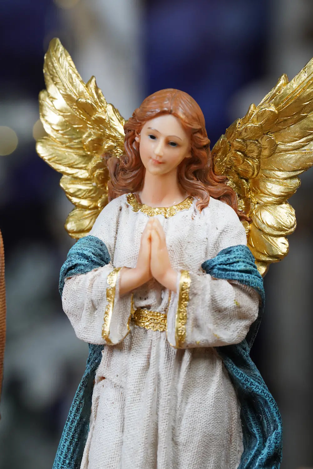 Nativity angel close-up — praying angel figurine with all-gold wings and white robe with teal sash against bokeh background