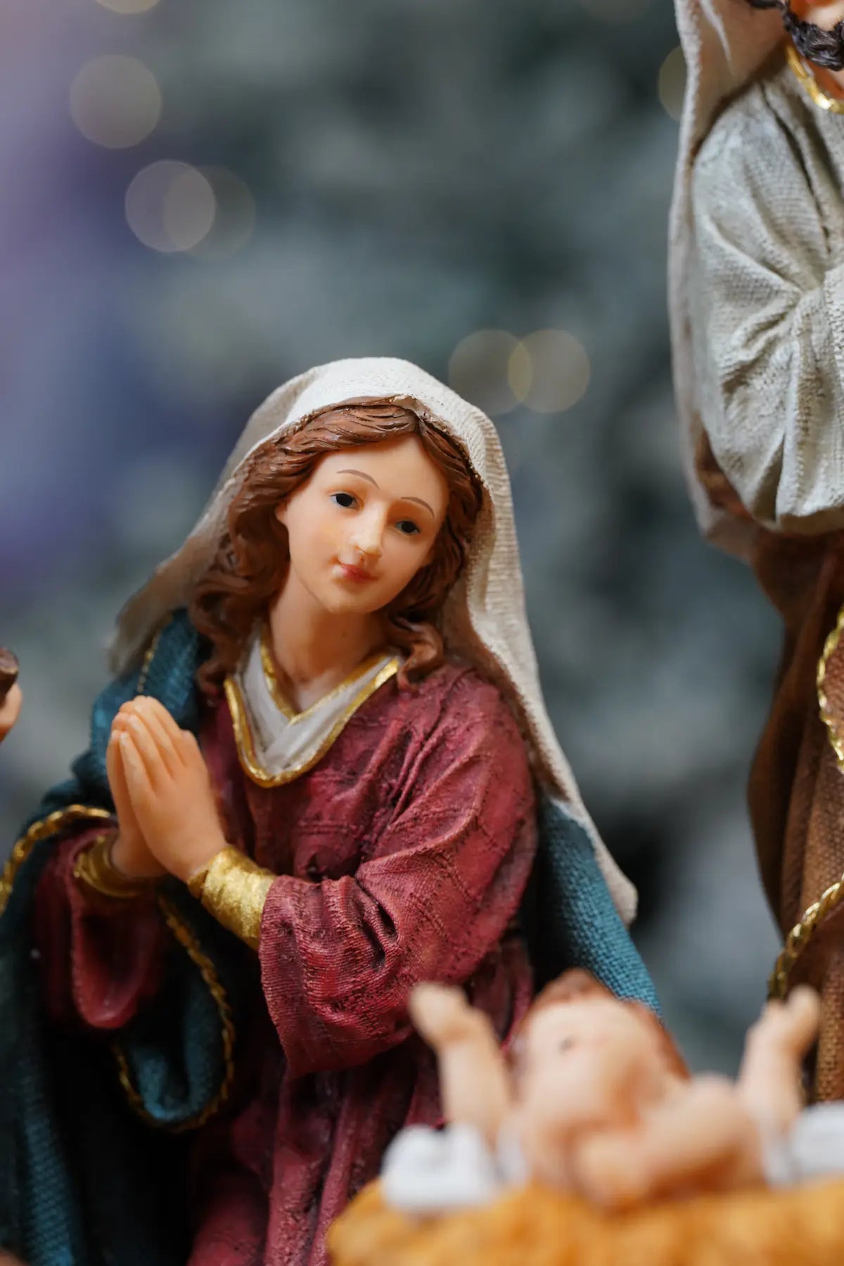 Virgin Mary nativity figurine close-up — kneeling Madonna in deep crimson robe with teal mantle and gold bracelet beside Baby Jesus manger