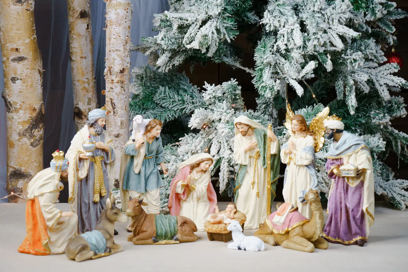 A Christmas nativity scene figurine set featuring Mary, Joseph, baby Jesus, three wise men, animals, and angels, arranged in front of a flocked Christmas tree and birch trees.