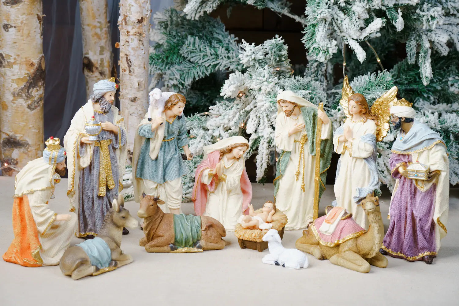 A Christmas nativity scene display featuring multiple hand-painted figurines of the Holy Family, wise men, angels, animals, and a manger, arranged in front of a flocked Christmas tree.