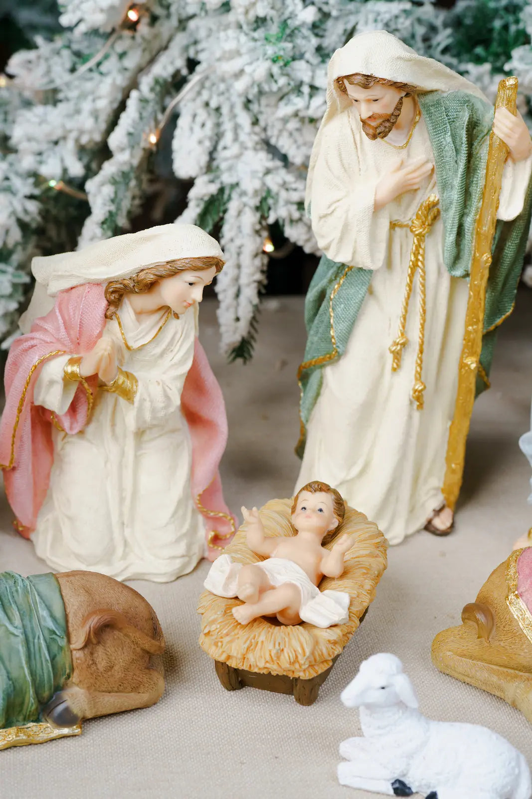 A Christmas nativity scene figurine set featuring Mary, Joseph, baby Jesus in a manger, a sheep, and a donkey, with a snowy Christmas tree in the background.