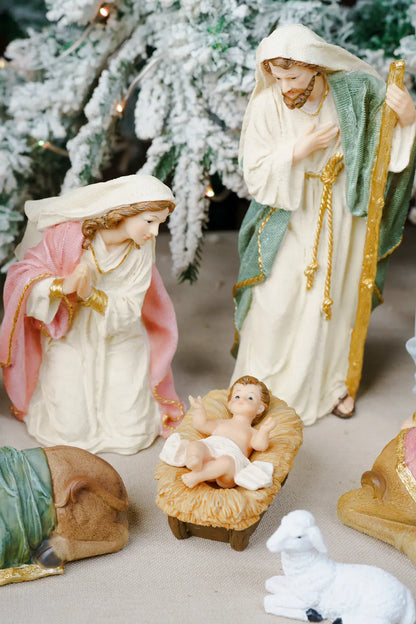 A Christmas nativity scene figurine set featuring Mary, Joseph, baby Jesus in a manger, a sheep, and a donkey, with a snowy Christmas tree in the background.