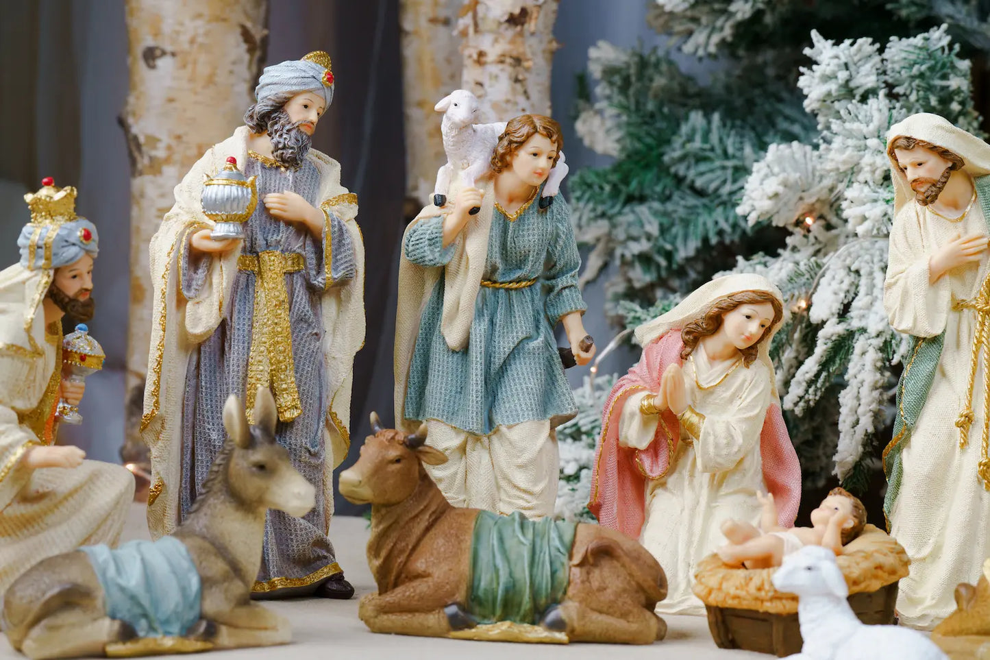 A Christmas nativity scene display featuring multiple painted figurines including Mary, Joseph, baby Jesus, wise men, an angel, a deer, and a cow, arranged in front of a decorated tree.