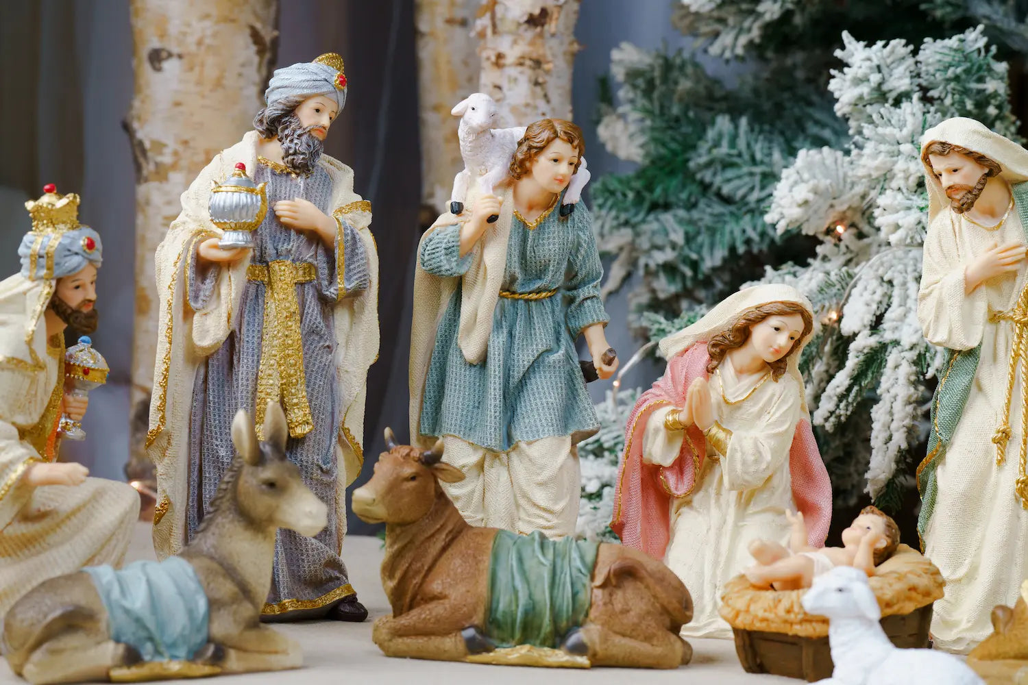 A Christmas nativity scene display featuring multiple painted figurines including Mary, Joseph, baby Jesus, wise men, an angel, a deer, and a cow, arranged in front of a decorated tree.