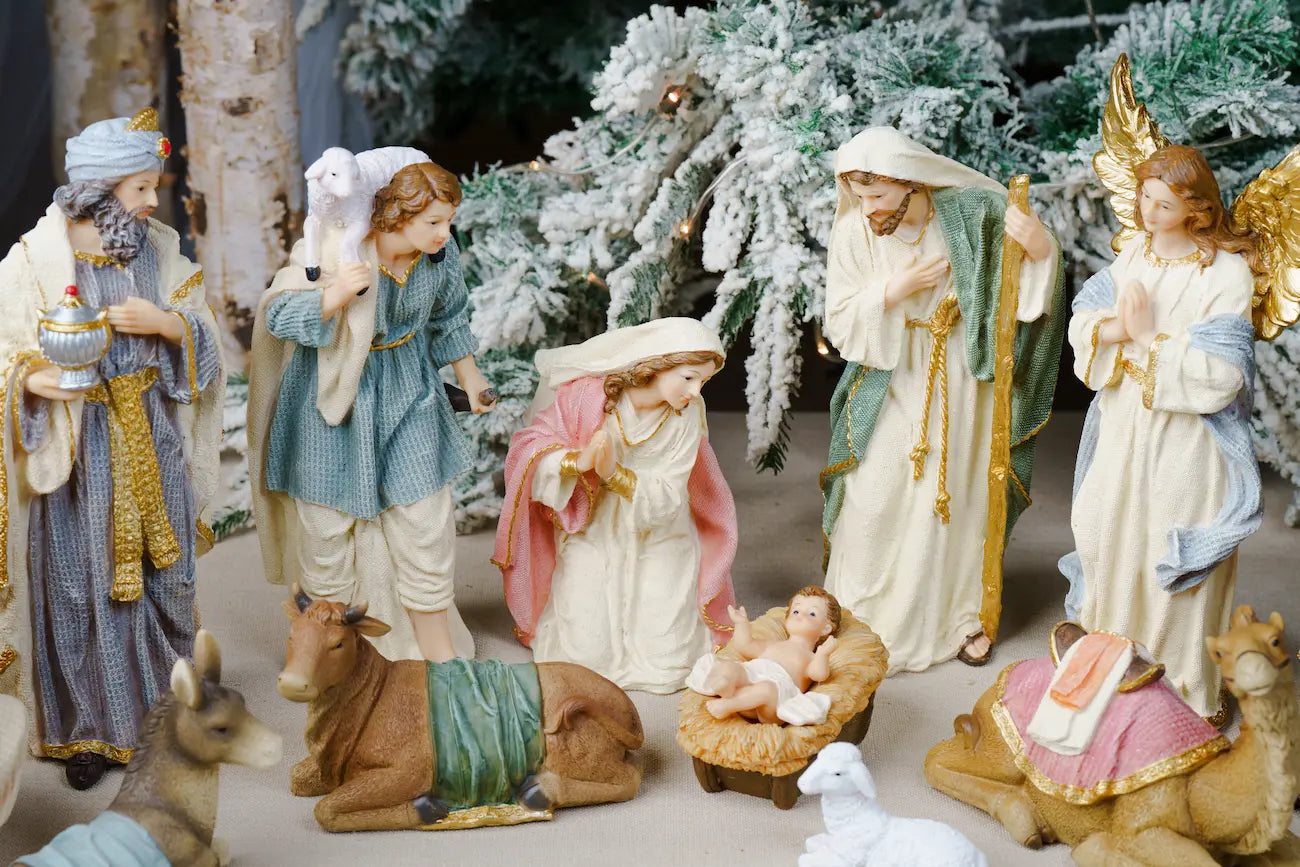 A Christmas nativity scene figurine set featuring Mary, Joseph, baby Jesus in a manger, wise men, angels, and animals, styled with frosted pine trees in the background.