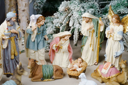 A Christmas nativity scene figurine set featuring Mary, Joseph, baby Jesus in a manger, wise men, angels, and animals, styled with frosted pine trees in the background.