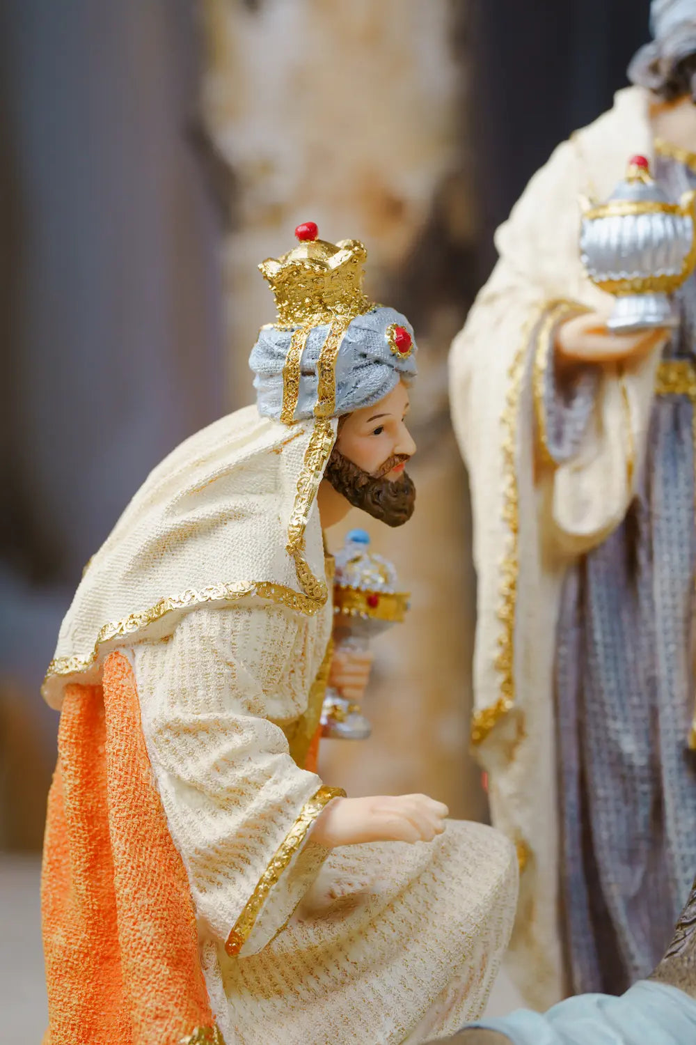 a kneeling king figurine in ornate cream robes with gold trim, orange sash, blue-gray turban, gold crown, and blurred robed figure in background.