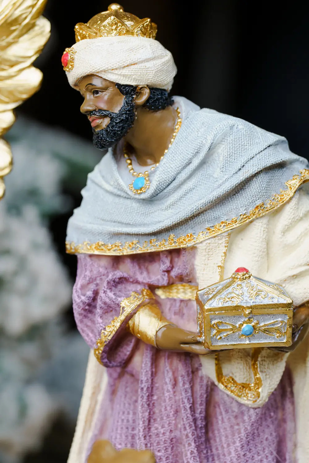 A wise man figurine or statue in regal attire, wearing a white turban, lavender-purple robe, and light gray shawl with gold trim, while holding a small ornate box.