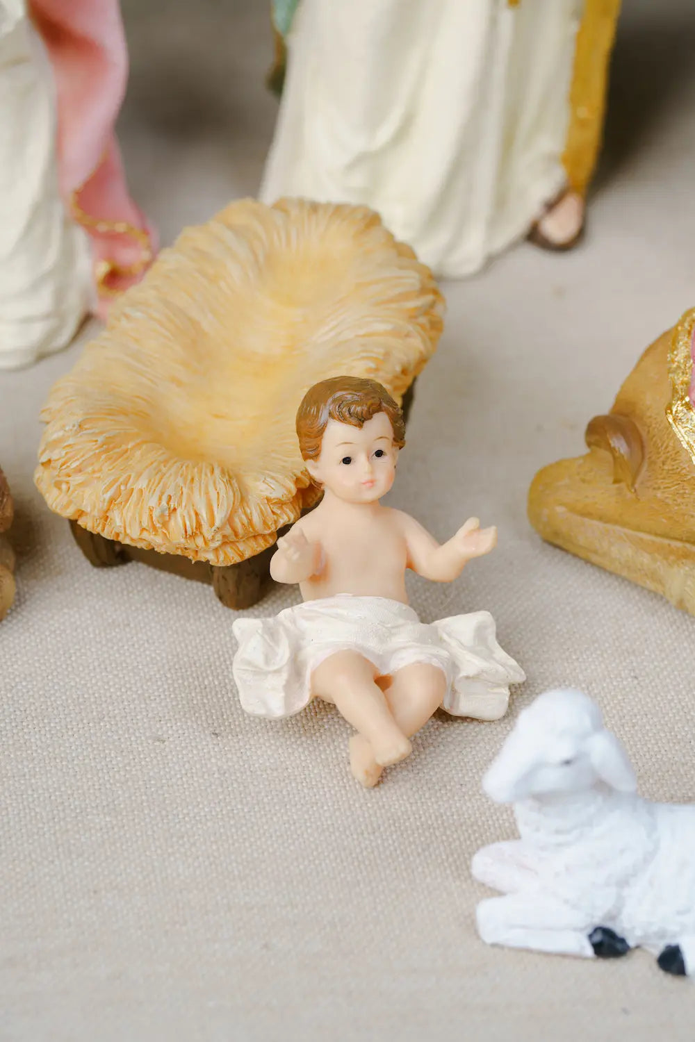 A baby Jesus figurine, placed in a nativity scene with a golden manger, a white sheep, and saint figurines on a beige fabric surface.