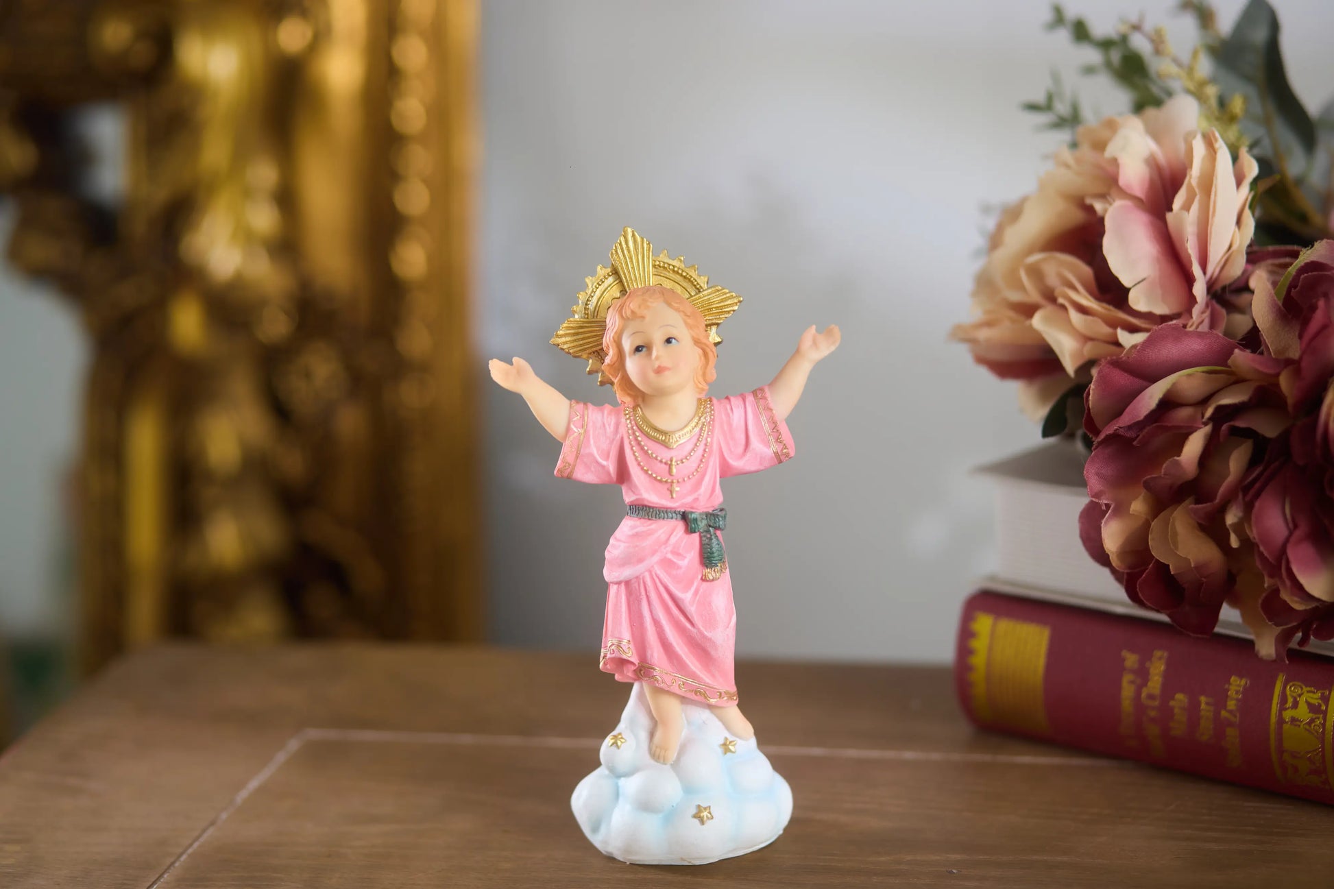 Niño Dios statue displayed on wooden table with golden frame background and floral decor