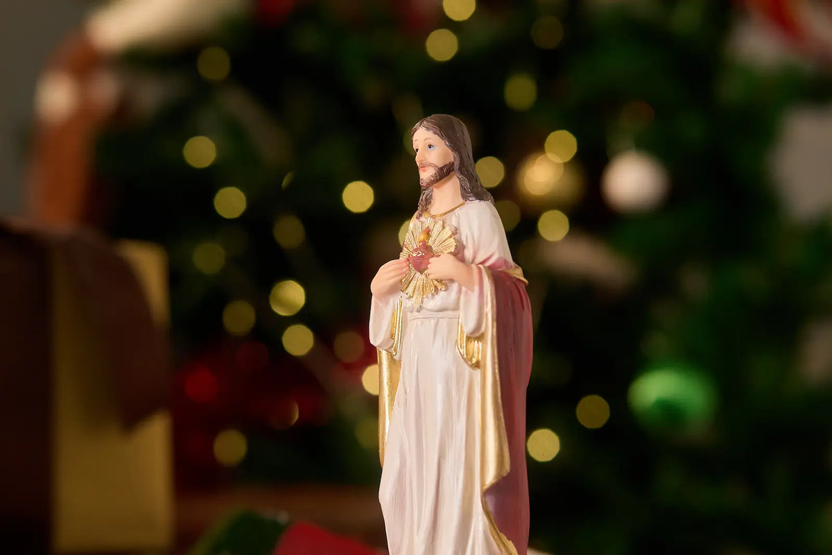 A figurine of the Sacred Heart of Jesus, dressed in a white robe with gold trim and a pinkish-red cloak, standing in front of a blurred Christmas tree with warm bokeh lights.
