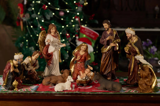Nativity scene displayed on red runner with Christmas tree lights, indoor tabletop holiday decor.