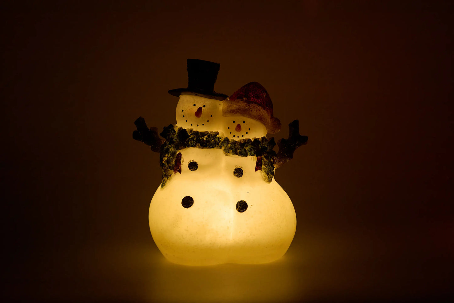 Snowman couple figurine glowing full warm amber in dark room — LED glow-through resin Christmas nightlight effect on black background