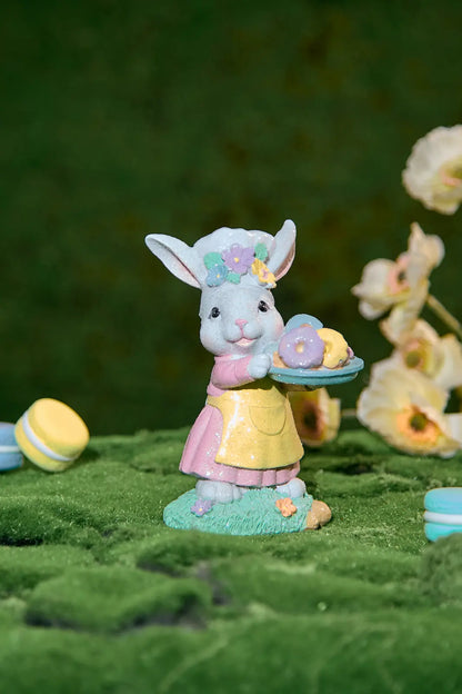 Close-up of Easter bunny chef figurine holding pastel donut tray — glitter resin spring decoration with flower toque and yellow apron on moss