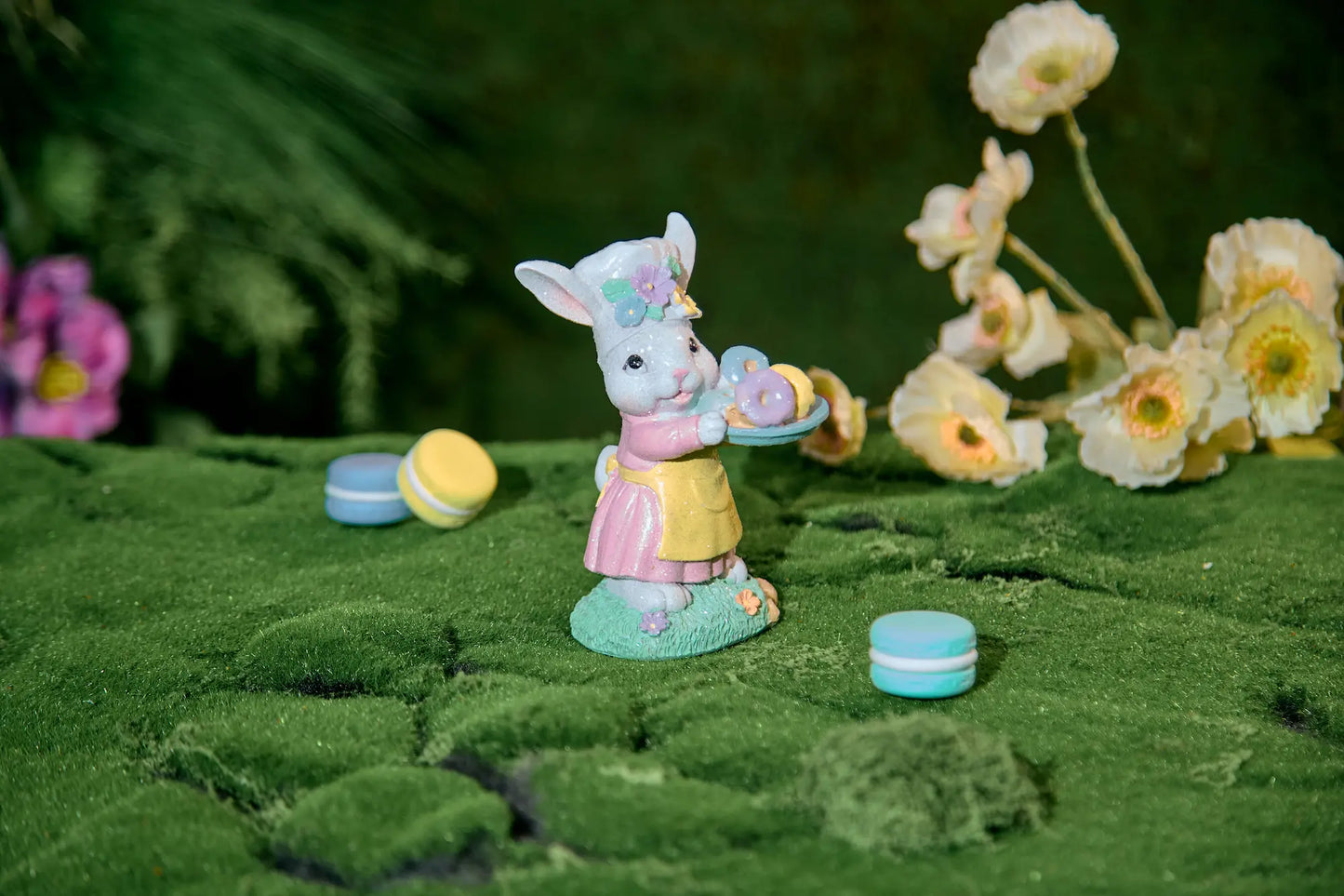 Easter bunny figurine with donut tray on moss surface — hand-painted glitter resin spring tabletop accent with bokeh flower background