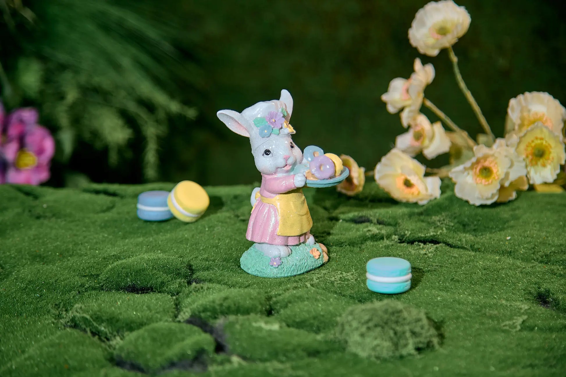 Easter bunny figurine with donut tray on moss surface — hand-painted glitter resin spring tabletop accent with bokeh flower background