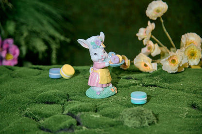 Easter bunny figurine with donut tray on moss surface — hand-painted glitter resin spring tabletop accent with bokeh flower background
