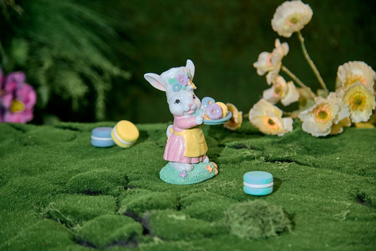 Easter bunny figurine with donut tray on moss surface — hand-painted glitter resin spring tabletop accent with bokeh flower background