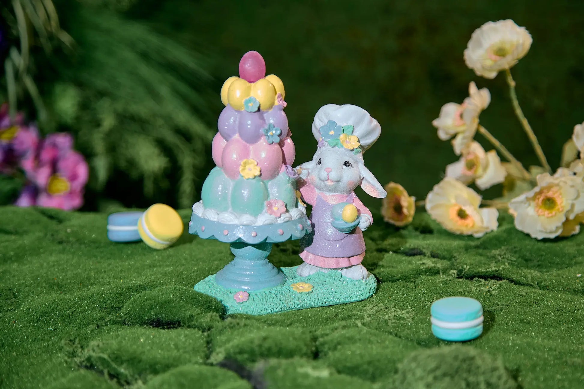 Pastel Easter bunny baker figurine on moss surface with spring flowers and macarons — glitter resin egg tower tabletop decoration in garden setting