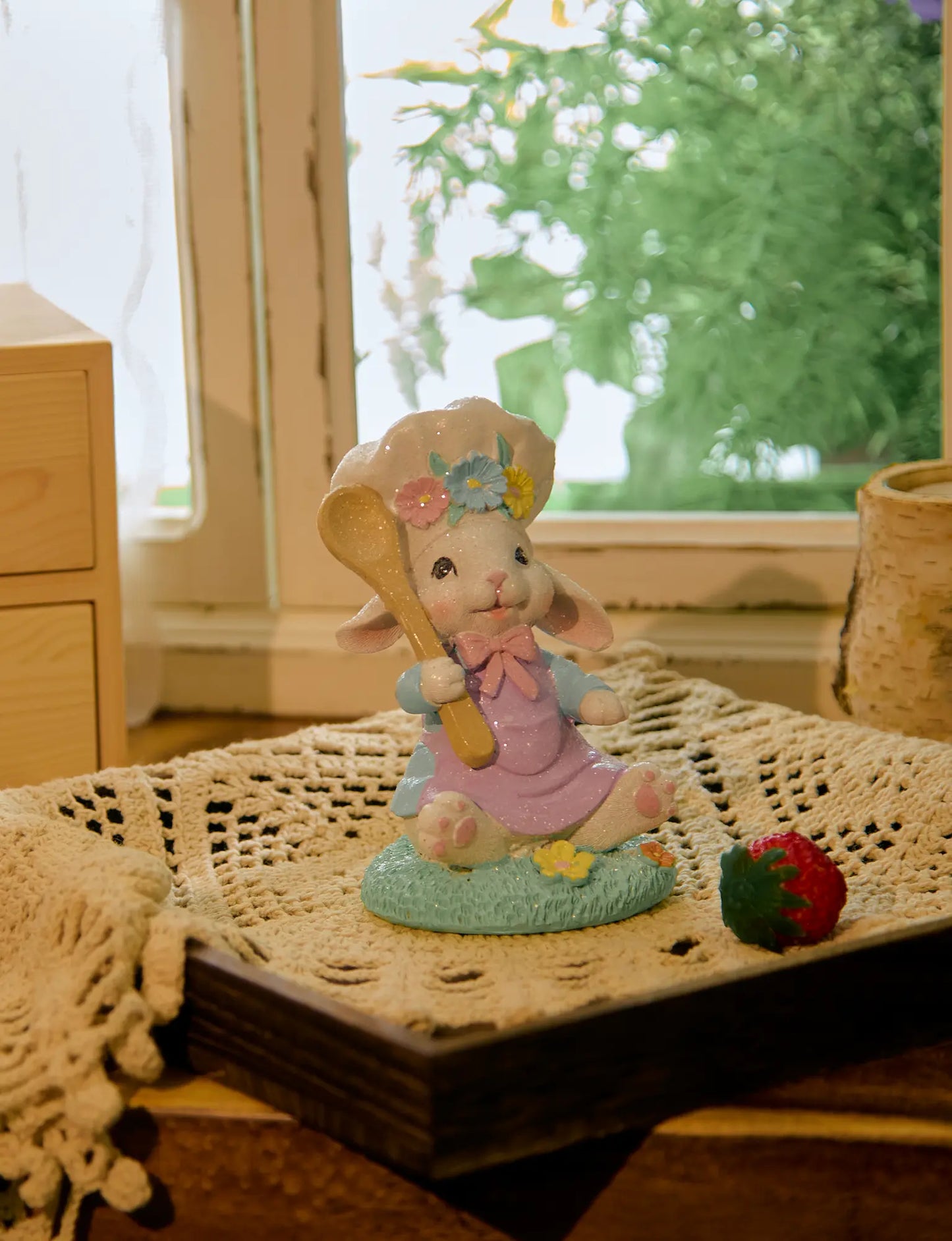 Seated Easter bunny chef figurine on crochet doily with strawberry beside cottage window — pastel glitter resin spring shelf sitter