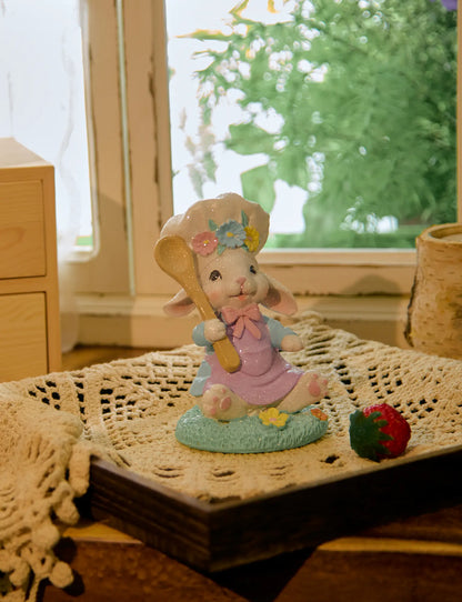 Seated Easter bunny chef figurine on crochet doily with strawberry beside cottage window — pastel glitter resin spring shelf sitter