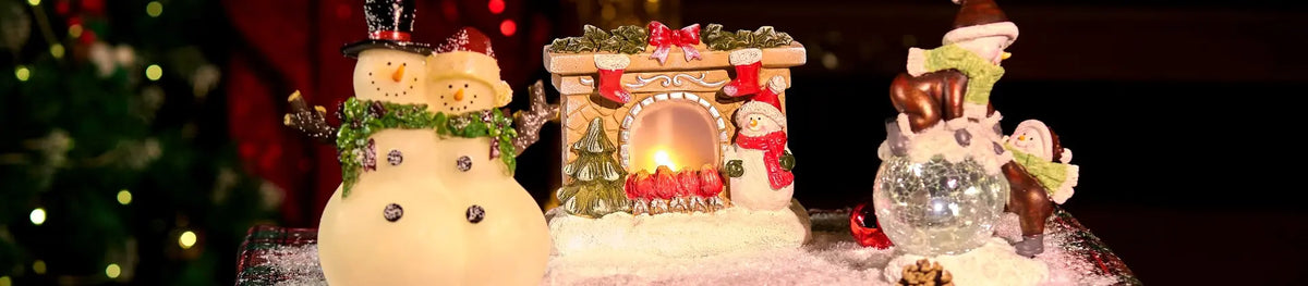 A festive Christmas tabletop display featuring a snowman ornament, a miniature glowing fireplace figurine, and a snow globe figurine, set against a dark blurred holiday background.