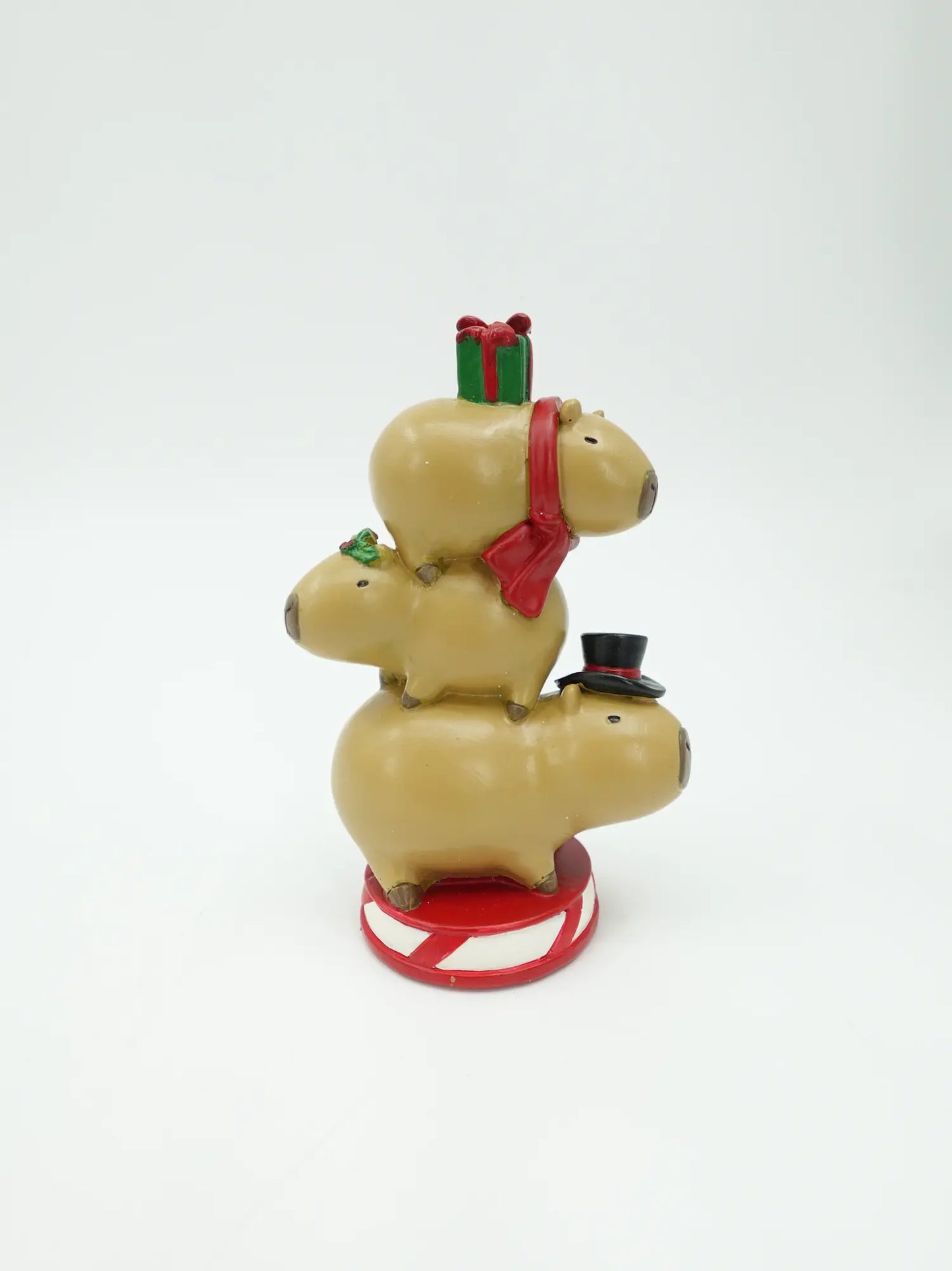 A festive capybara figurine stacked in three sizes, decorated with a red scarf, a small green gift box on top, a black top hat, and a red-and-white striped base.