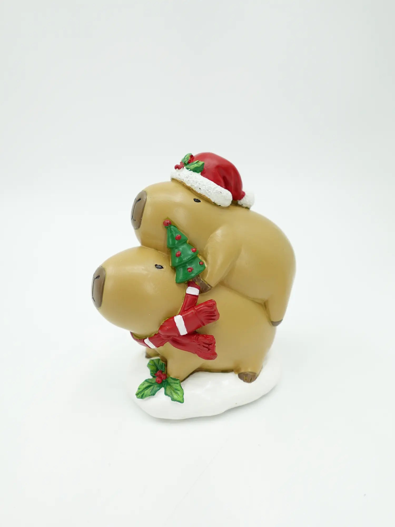 A holiday figurine of two stacked capybara on a white base, decorated with a red Santa hat, a green Christmas tree