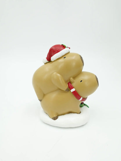 A capybara figurine in tan, wearing a red Santa hat and a red holiday collar, standing on a white base against a plain white background.